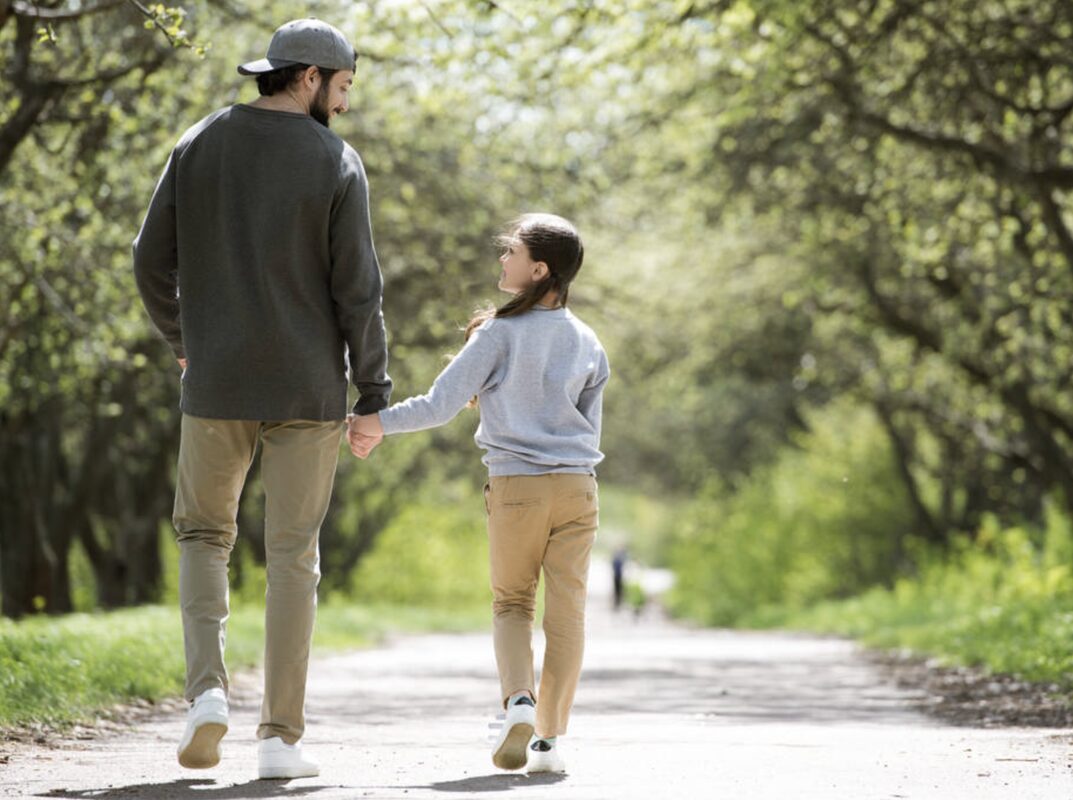 A father and his daughter walking hand in hand down a tree-lined park path